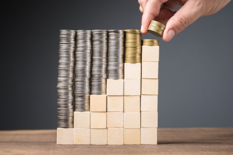 A hand is stacking gold coins on top of wooden blocks arranged in ascending order, next to stacks of silver coins on other wooden blocks. The arrangement symbolizes financial growth or investment progress against a dark background.