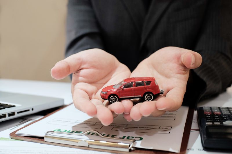 Person in a suit holding a small red toy car in their open hands over a clipboard with a car insurance form, next to a laptop and calculator on a desk, symbolizing car insurance protection and planning.