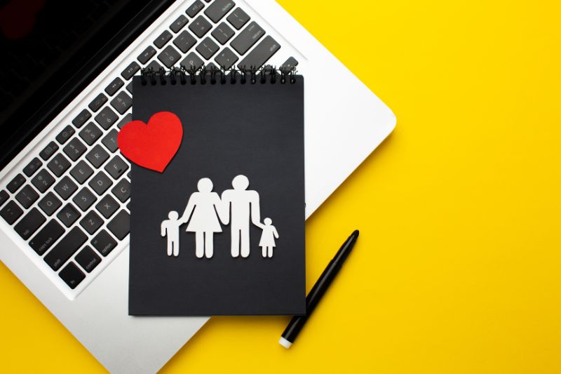 Black notepad with cut-out paper family and red heart placed on a laptop keyboard with yellow background, symbolizing family protection and insurance