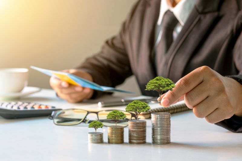 Business person in a suit arranging stacked coins with small trees growing on top, symbolizing financial growth and investment.