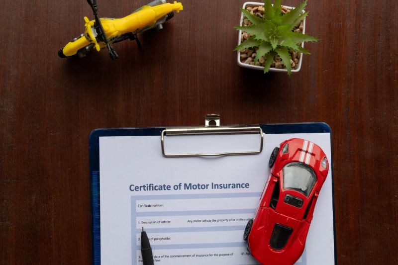 Certificate of motor insurance document on a clipboard with a red toy car, yellow toy bike, pen, and small potted plant placed on a wooden table.