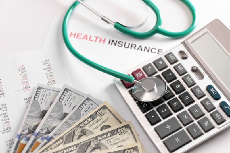 A stethoscope resting on a calculator beside a document labeled “Health Insurance,” with U.S. dollar bills and financial charts on a desk.