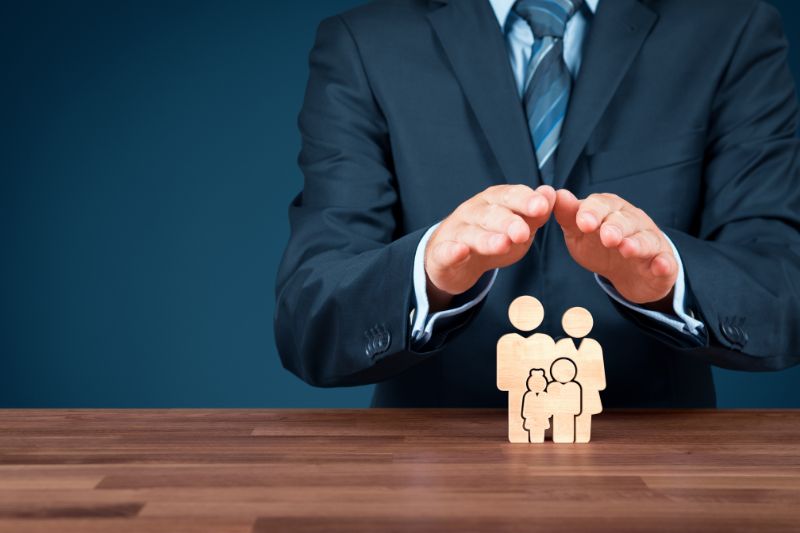 A person in a business suit holds their hands protectively over small wooden family-shaped figures on a table, symbolizing insurance, safety, or family protection.