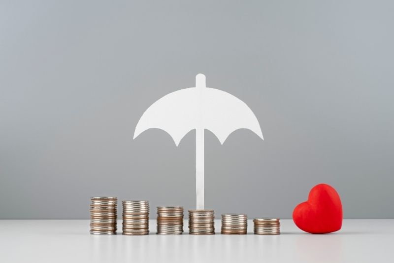 Stacks of coins under a white umbrella icon symbolizing financial protection, with a red heart placed beside them on a neutral background.