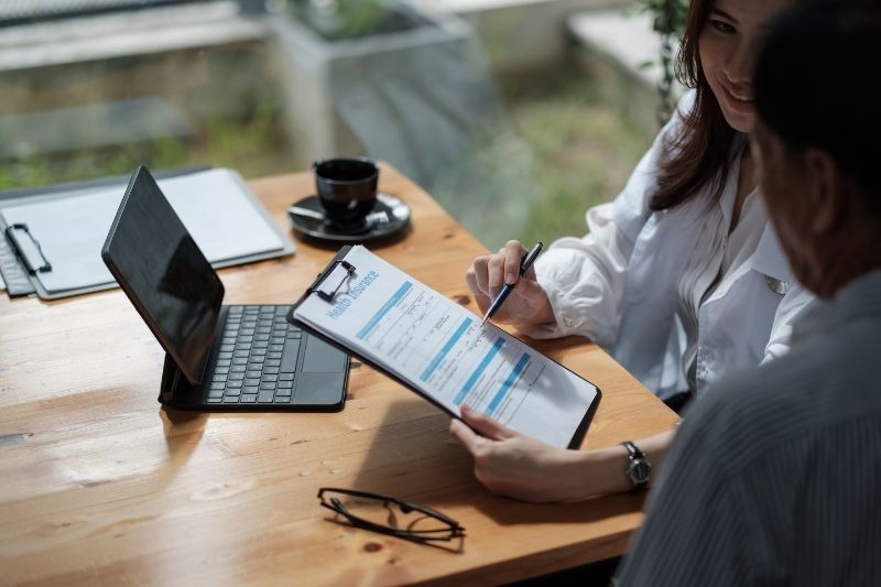 Two people seated at a wooden table reviewing a document on a clipboard, with a laptop, coffee cup, and eyeglasses nearby, appearing to discuss paperwork or a form.