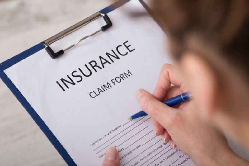 Person holding a pen and filling out an insurance claim form on a clipboard, with the word “INSURANCE” prominently displayed at the top.