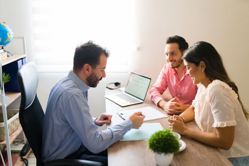 A financial advisor sits at a desk in a bright office, reviewing documents with a smiling young couple. A laptop and paperwork are on the table, and a small potted plant is in the foreground.
